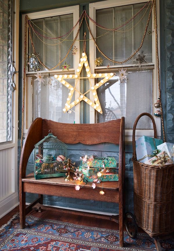 Old wooden bench with wrapped packages and birdcage filled with ornaments. Wicker shopping cart and star decorations on porch.