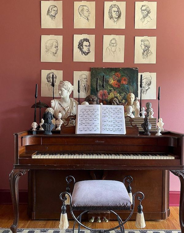 Old piano with sketches taped to wall and vintage busts and candlesticks.